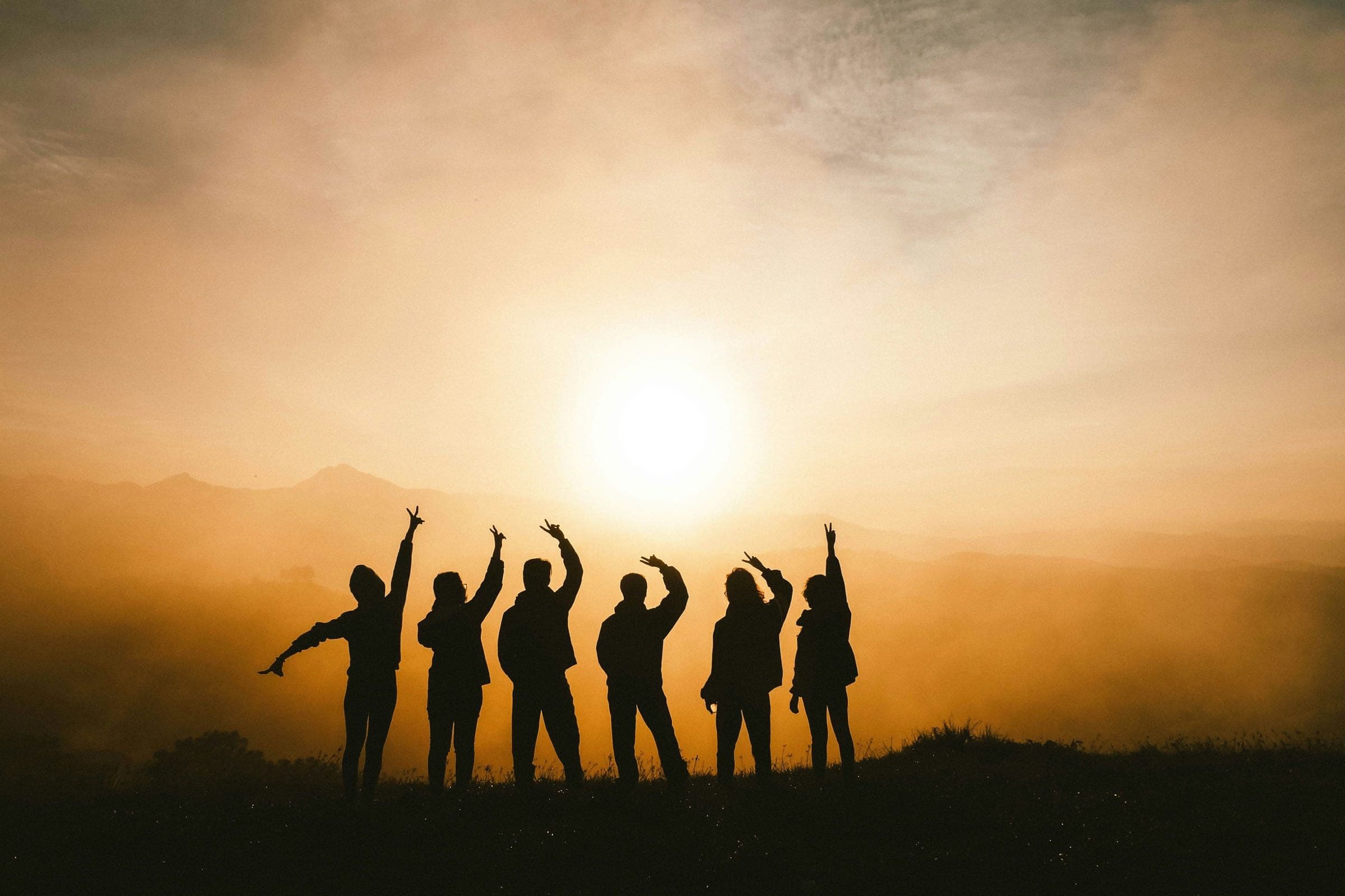 Six friends silhouetted on a hilltop at sunrise, arms raised towards the sun