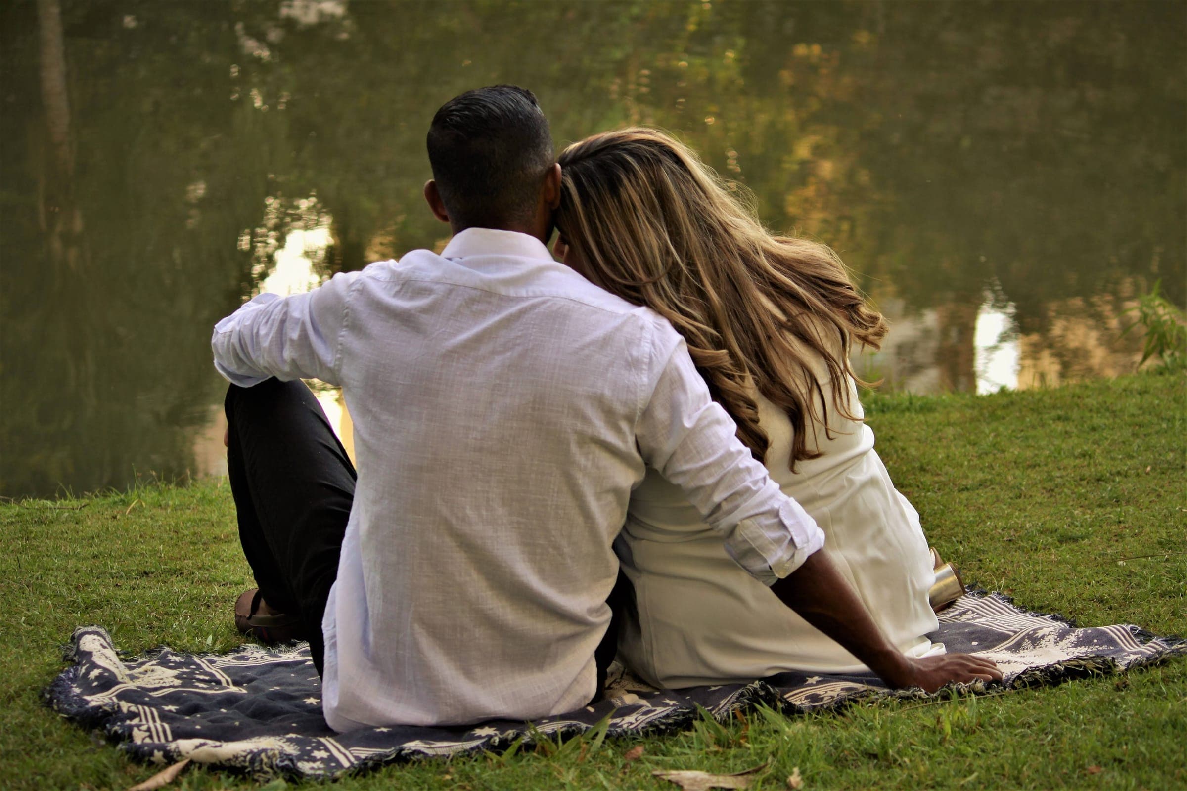 A couple sitting close together on a blanket by a lake, seen from behind