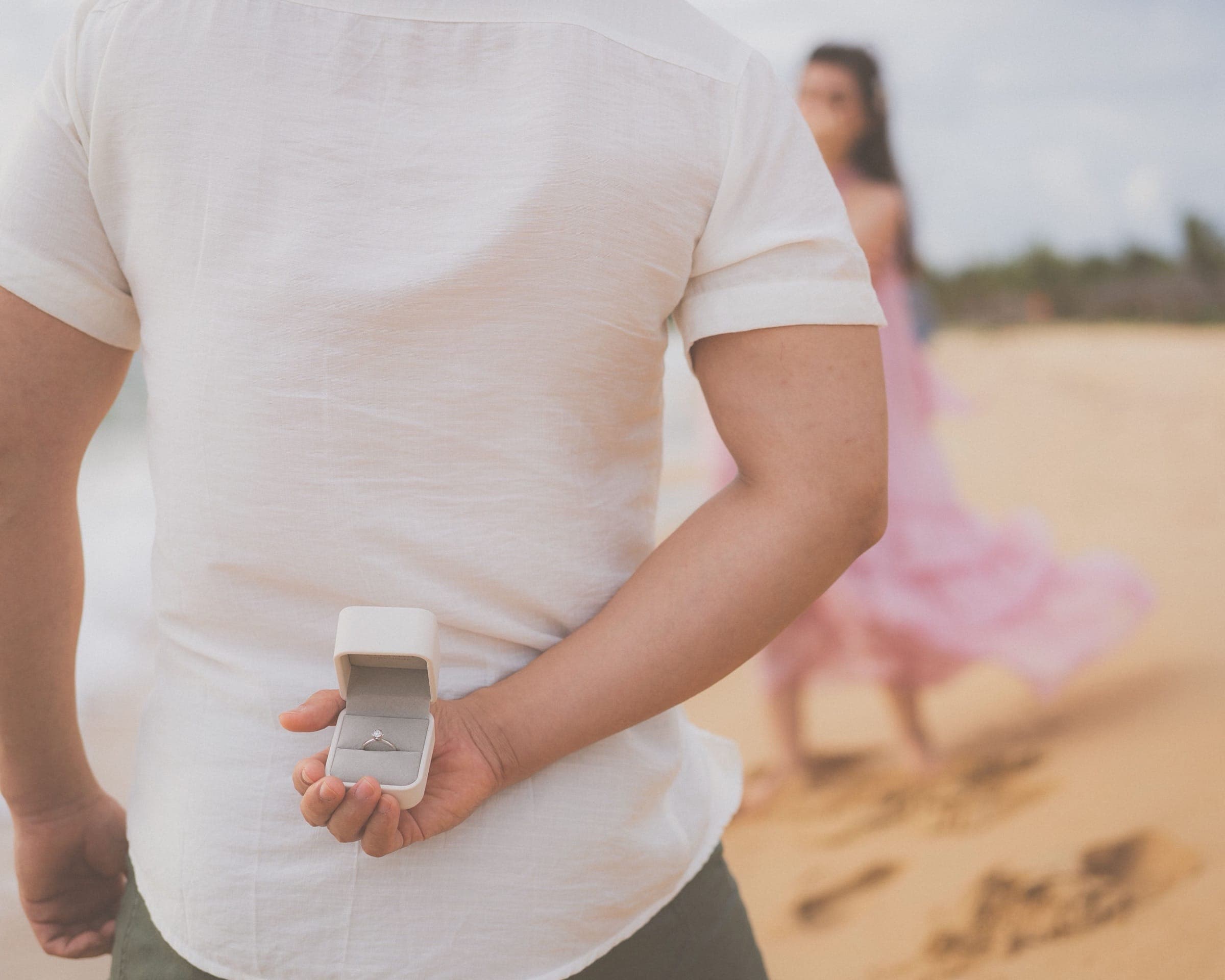 A man hides an engagement ring behind his back, preparing to propose on a beach
