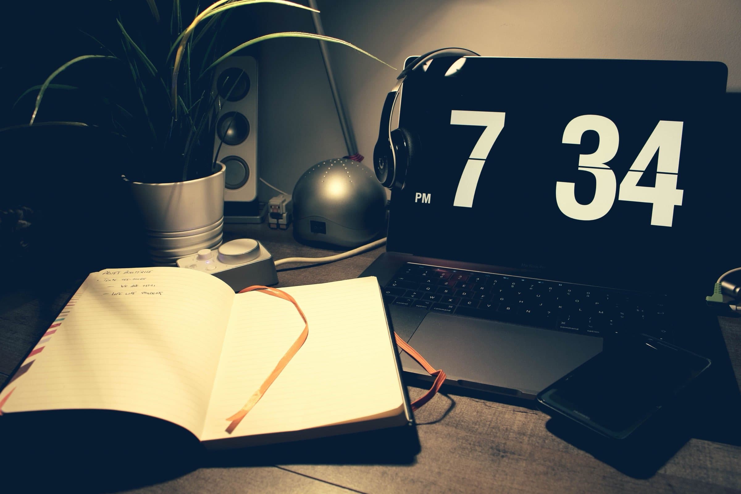 A laptop showing 7:34 PM beside an open notebook on a desk in the evening