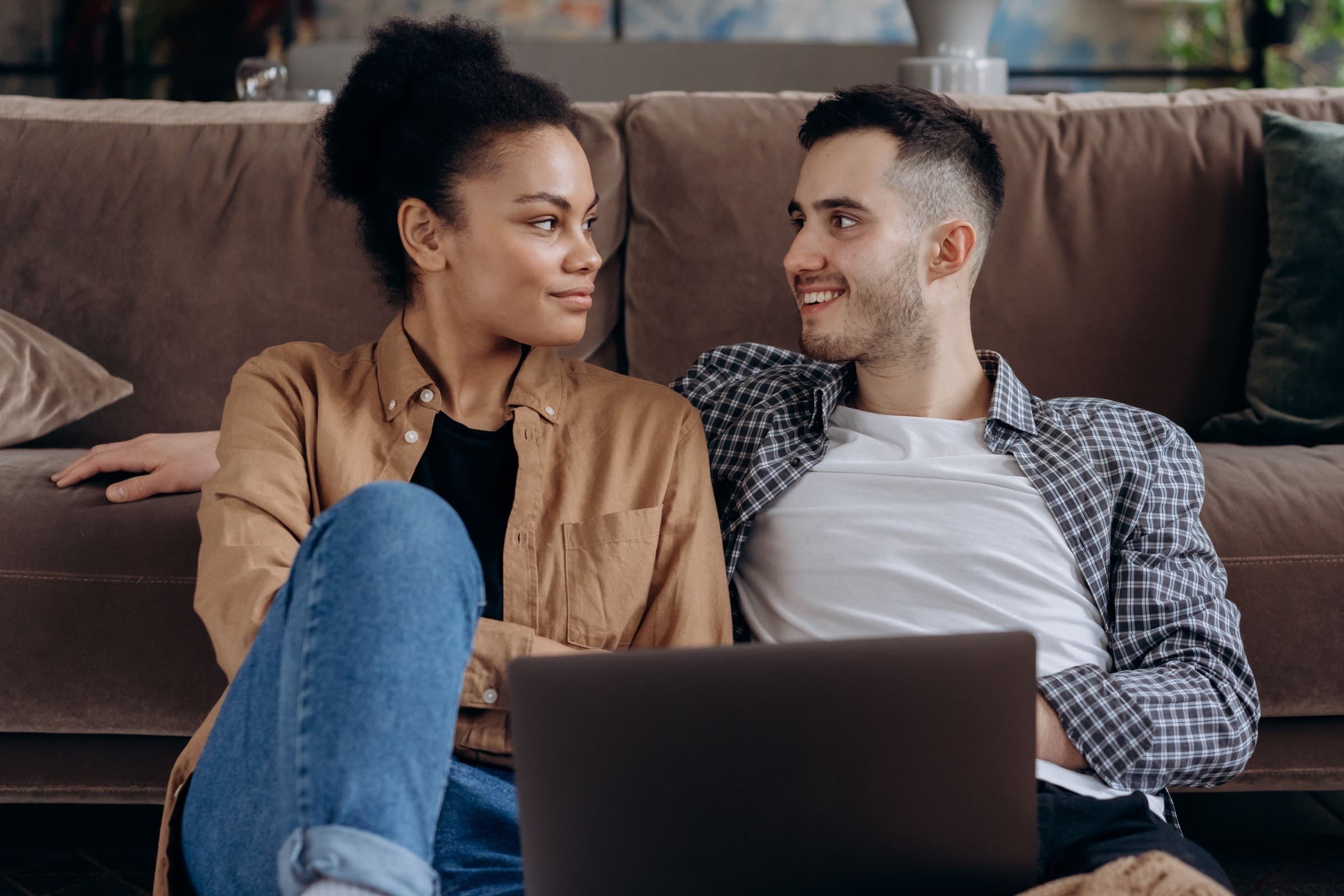 A smiling couple sitting together on the floor against a sofa, looking at each other over a laptop